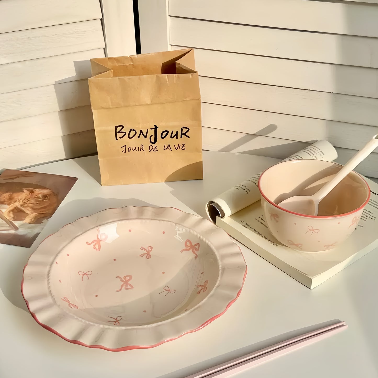 Table setting with a plate, bowl, and bag labeled 'Bonjour' on a light surface.