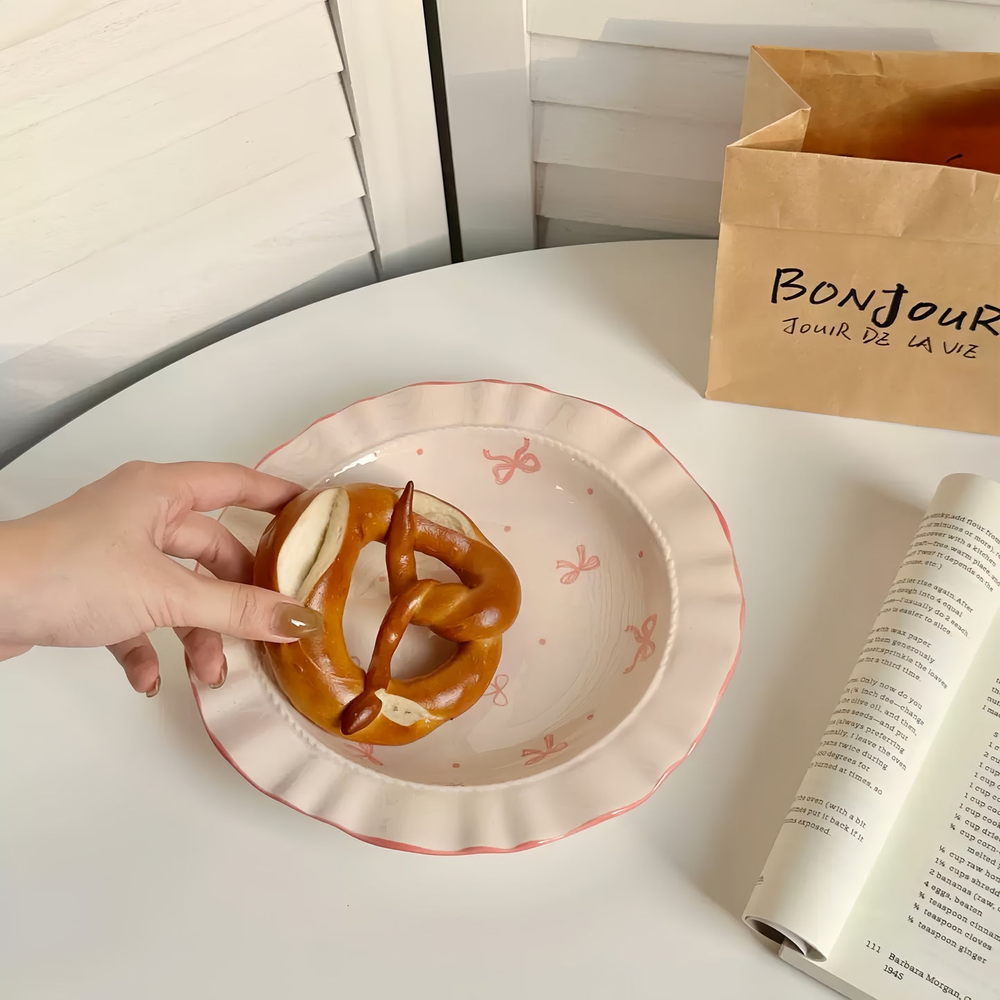 Person holding a pretzel over a decorative plate with a 'Bonjour' bag and open book in the background.