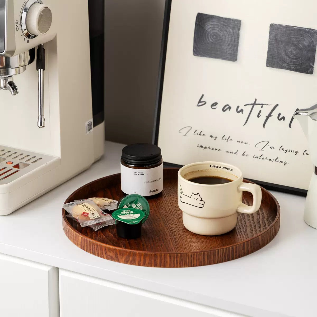 Café setting with a coffee machine, wooden tray with coffee cup and capsules, and framed quote on a white surface.